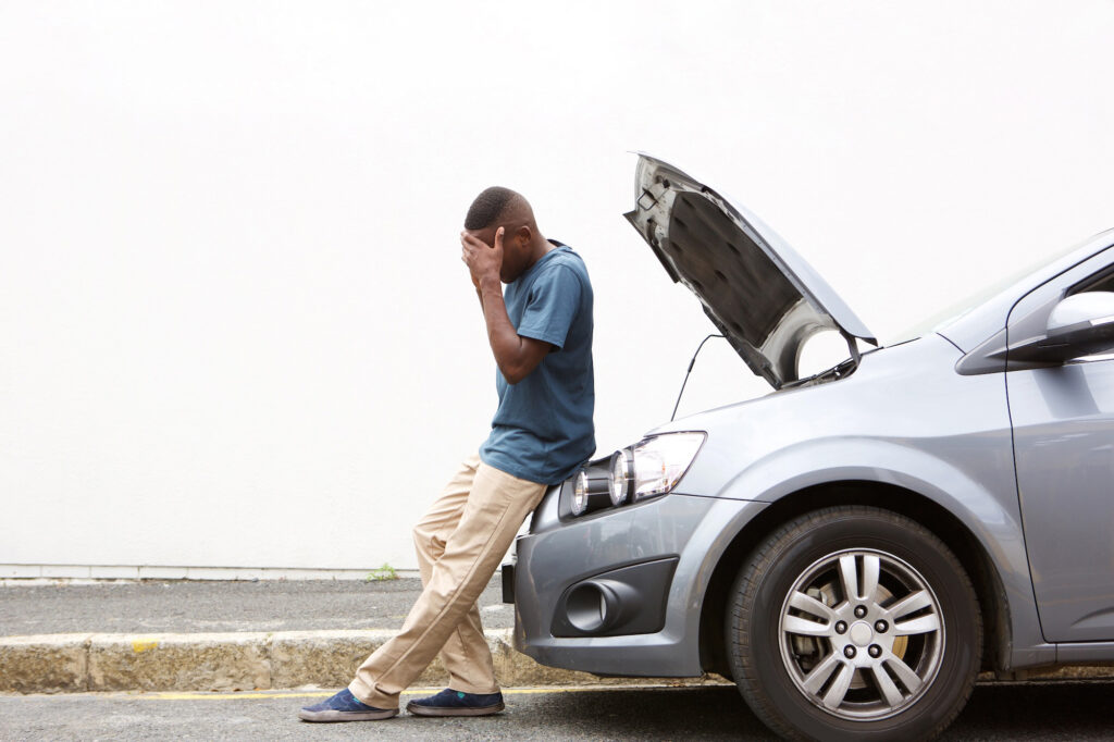 A man leans against his car with the hood open, covering his face in frustration as he deals with a breakdown due to a dead car battery.