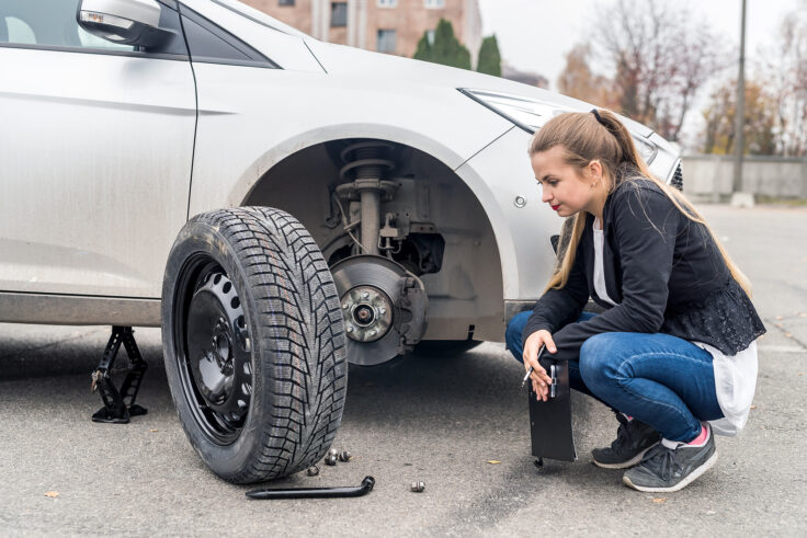 A woman crouches next to her car with the front tire removed, preparing to change it. The car is jacked up, and the tools and lug nuts are laid out on the ground in front of her as she inspects the wheel hub.