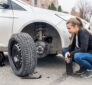 A woman crouches next to her car with the front tire removed, preparing to change it. The car is jacked up, and the tools and lug nuts are laid out on the ground in front of her as she inspects the wheel hub.