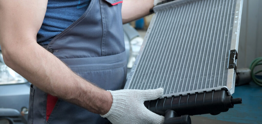 Mechanic holding a car radiator, wearing a work uniform and protective gloves, preparing for installation or inspection.