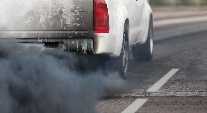 The rear view of a white pickup truck emitting thick black exhaust smoke onto the road, illustrating vehicle pollution and its impact on air quality.