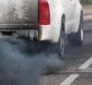 The rear view of a white pickup truck emitting thick black exhaust smoke onto the road, illustrating vehicle pollution and its impact on air quality.