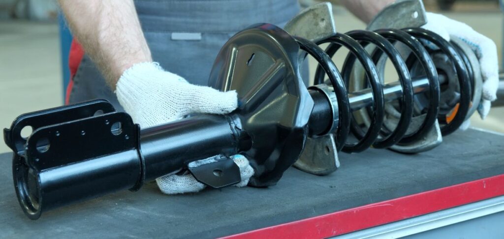 Mechanic wearing gloves holding a car strut assembly, including a coil spring and shock absorber, on a workbench.