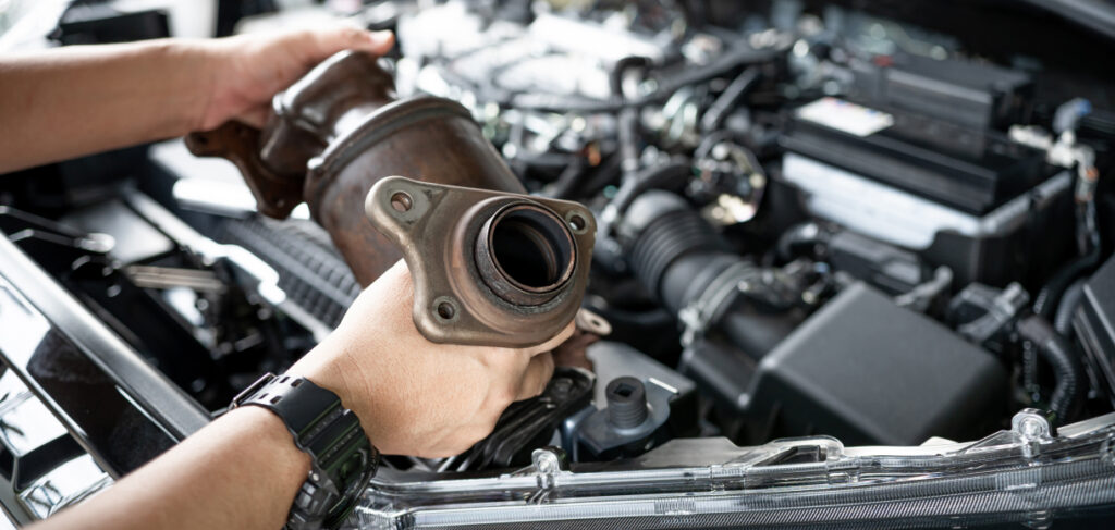 Mechanic holding a catalytic converter above an open car engine bay, inspecting or preparing for installation.