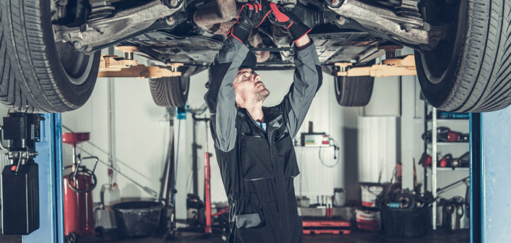 Mechanic in a workshop inspecting the underside of a car lifted on a hydraulic lift, wearing a black uniform and red gloves.