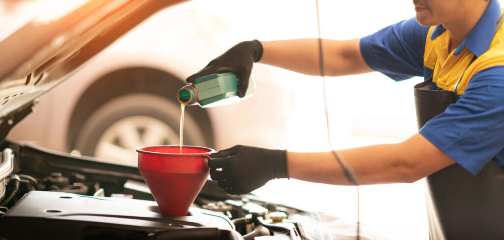 Mechanic in a blue and yellow uniform pouring engine oil into a car using a red funnel during a routine maintenance service.