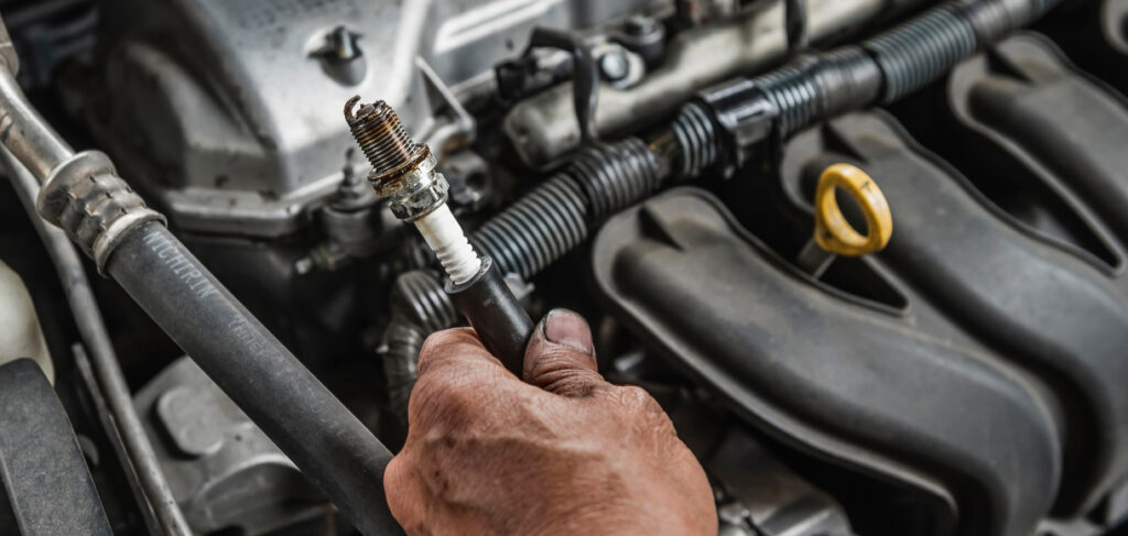 Mechanic holding a used spark plug while working on a car engine, with visible hoses, wiring, and engine components in the background.