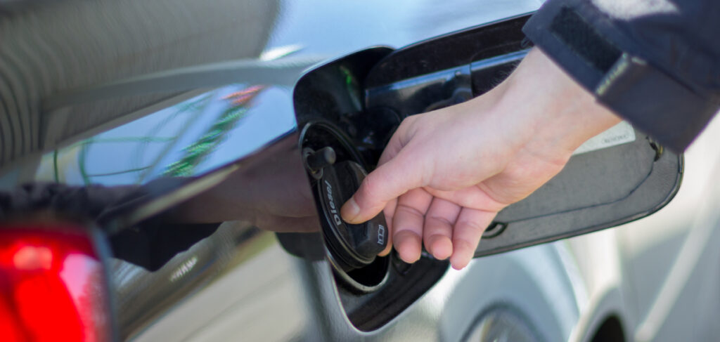 Person’s hand tightening a gas fuel cap on a car, with the fuel door open and the vehicle’s glossy exterior reflecting surroundings.