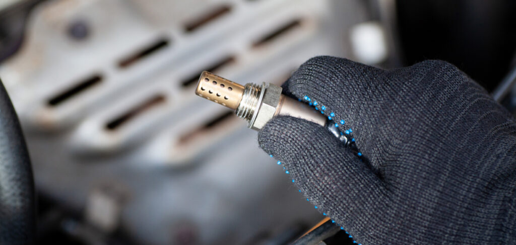 Mechanic wearing a black glove holding an oxygen sensor removed from a car engine, with metal components visible in the background.