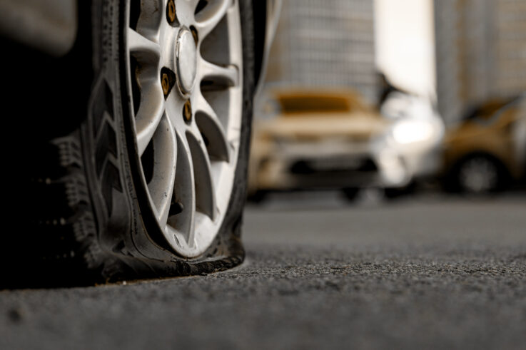 A close-up of a car's flat tire with the rim pressing against the ground, while other parked cars are blurred in the background.