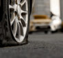 A close-up of a car's flat tire with the rim pressing against the ground, while other parked cars are blurred in the background.