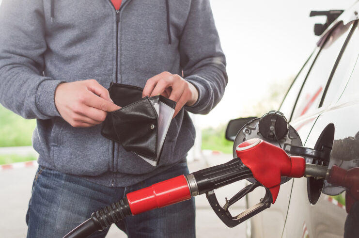 A person at a gas station holds an open wallet while fueling their car with a red gasoline nozzle, reflecting the financial strain of rising fuel costs.