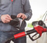 A person at a gas station holds an open wallet while fueling their car with a red gasoline nozzle, reflecting the financial strain of rising fuel costs.