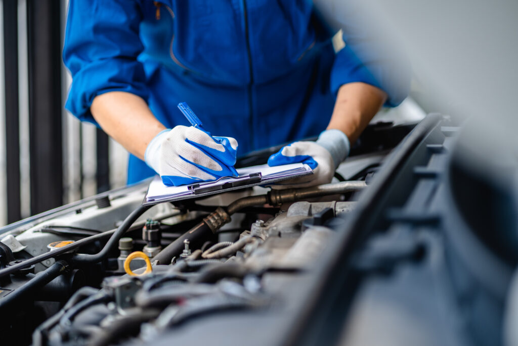 A mechanic wearing blue coveralls and gloves writes on a clipboard while inspecting a car engine, focusing on vehicle maintenance and repairs.