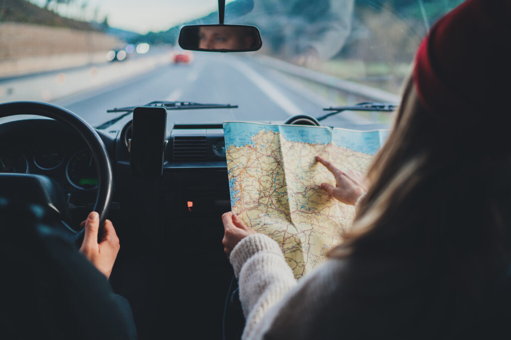 Two people in a car on a road trip, with the passenger holding and pointing at a paper map while the driver navigates down a highway.